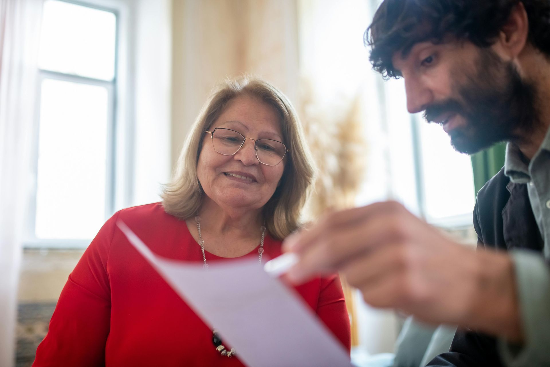 Woman in red shirt and glasses smiles while man points at paper in bright room.