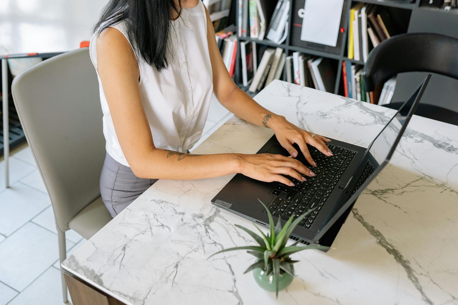 Woman typing on a laptop at a marble table, small plant nearby. Bookshelf visible.