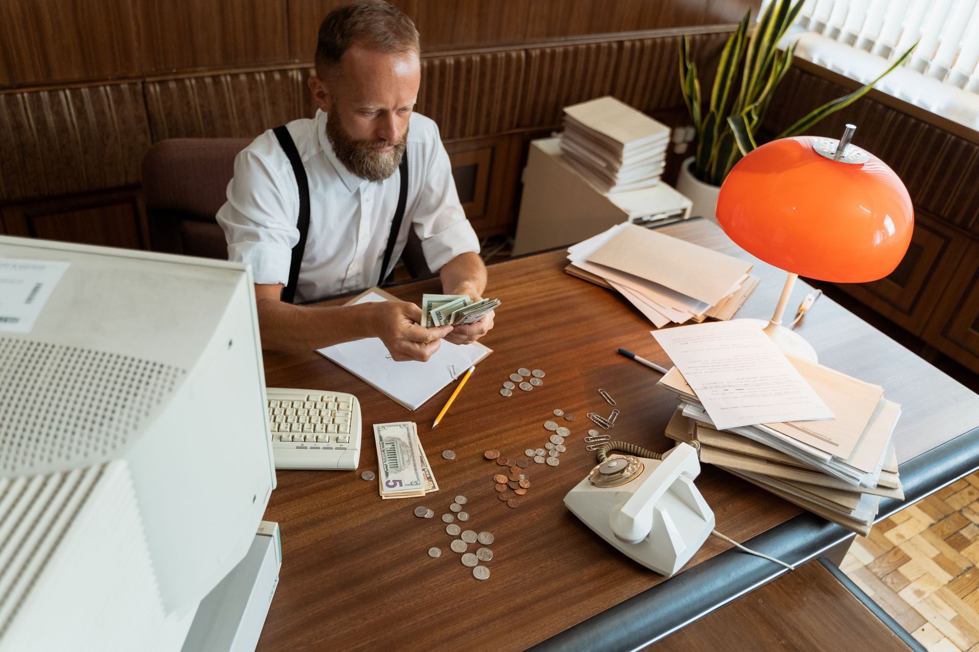 Man counting money at a cluttered desk with papers, calculator, and orange lamp