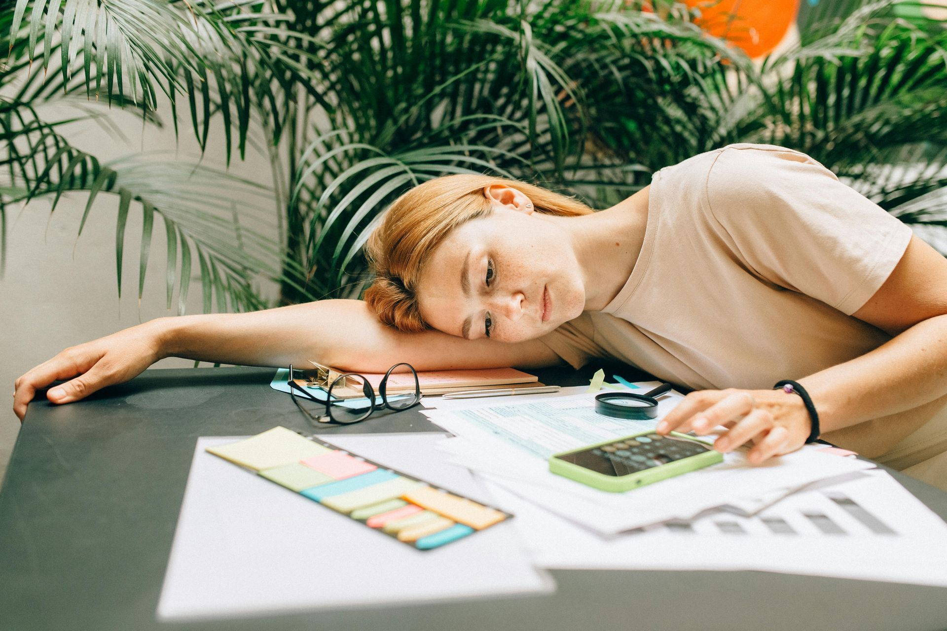 Person asleep at a desk with paperwork, calculator, and glasses in a bright office