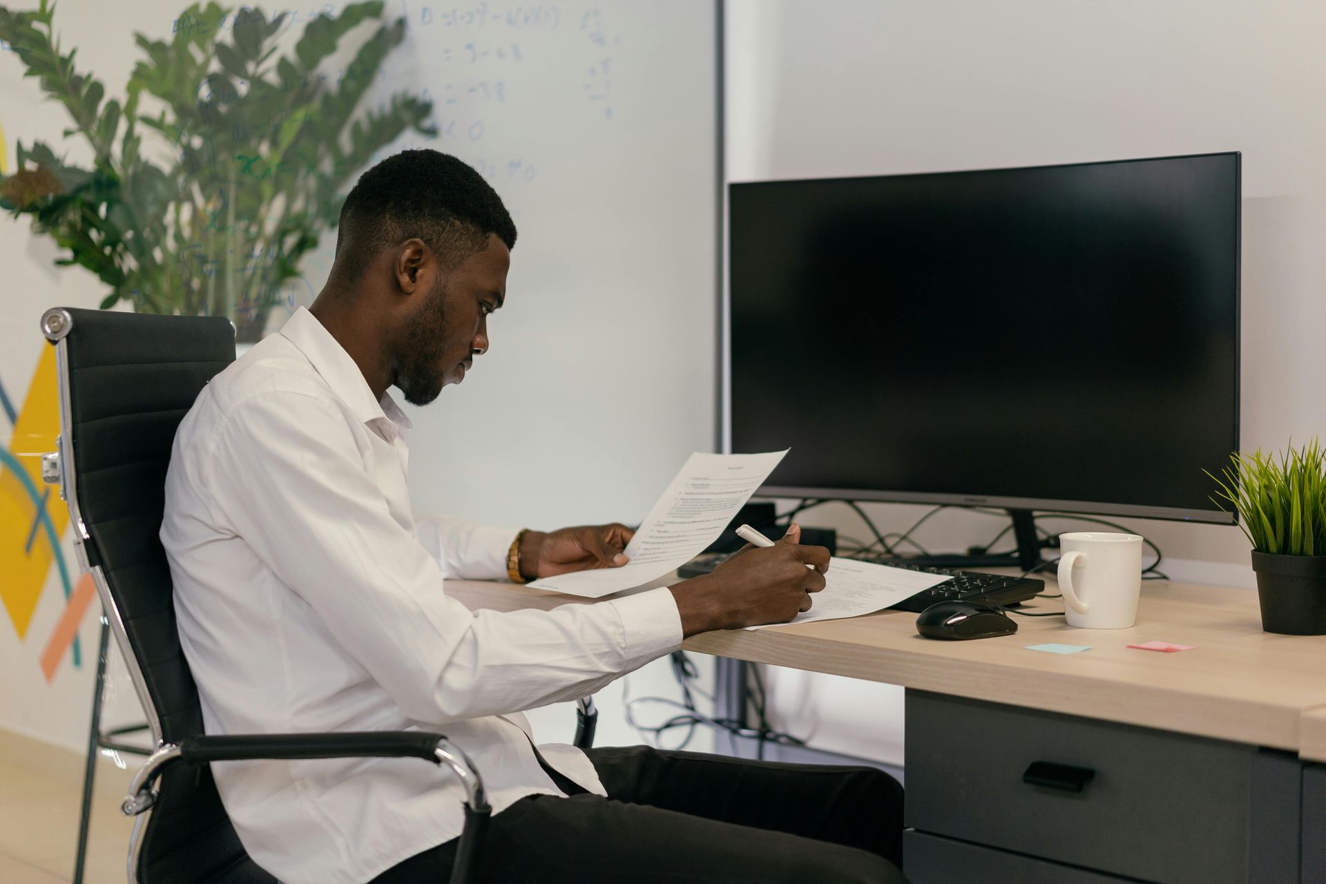 Man in white shirt sitting at a desk, reviewing papers, with a computer monitor in a modern office.