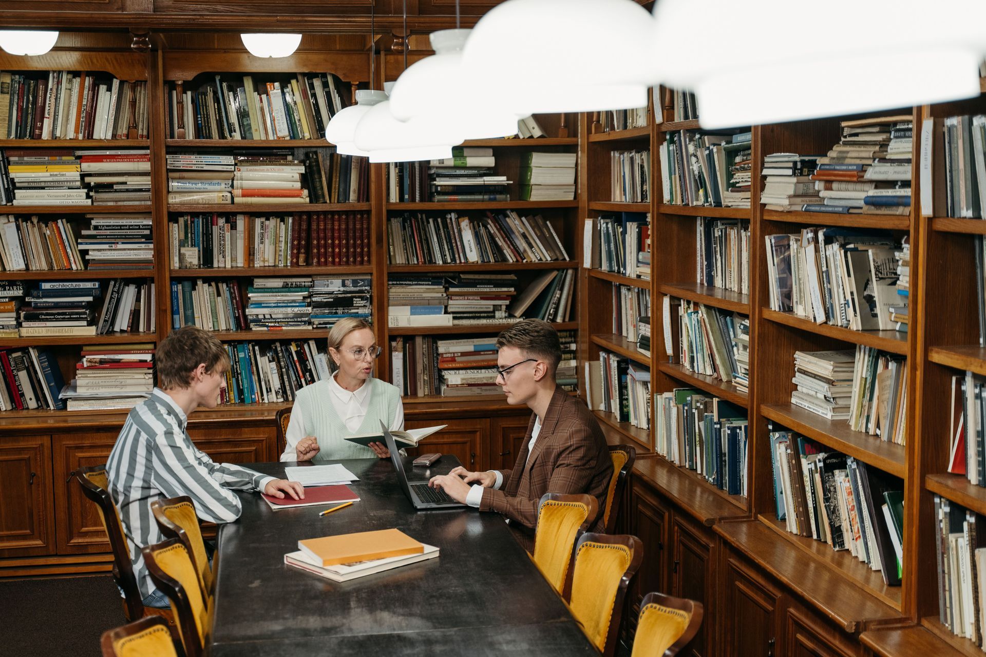 Three people sit at a table in a wood-paneled library, talking with bookshelves all around.