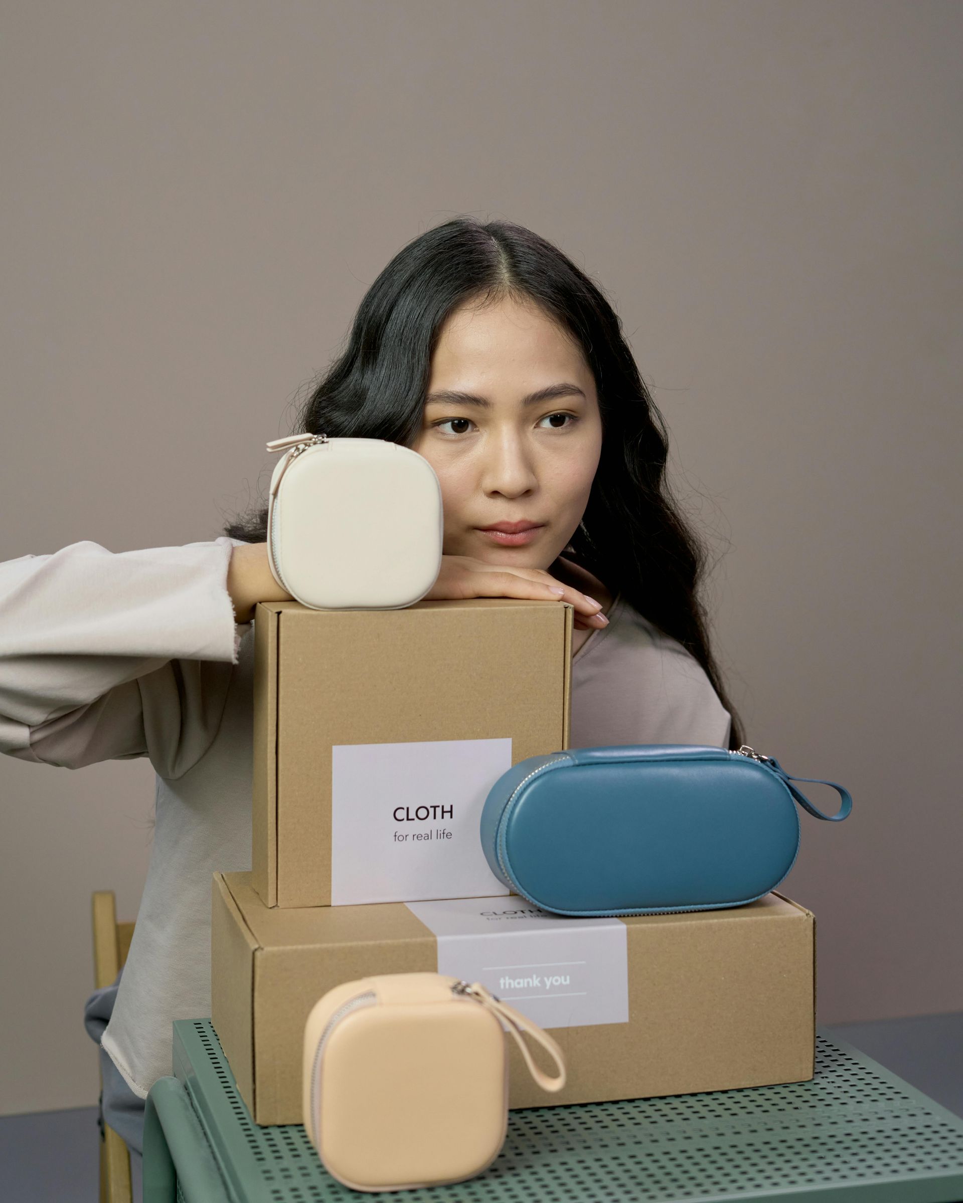 Woman leans on stacked boxes with three small pouches: cream, teal, and peach.
