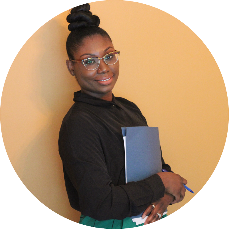 Woman with glasses, hair bun, holding file and pen, standing in front of an orange wall.