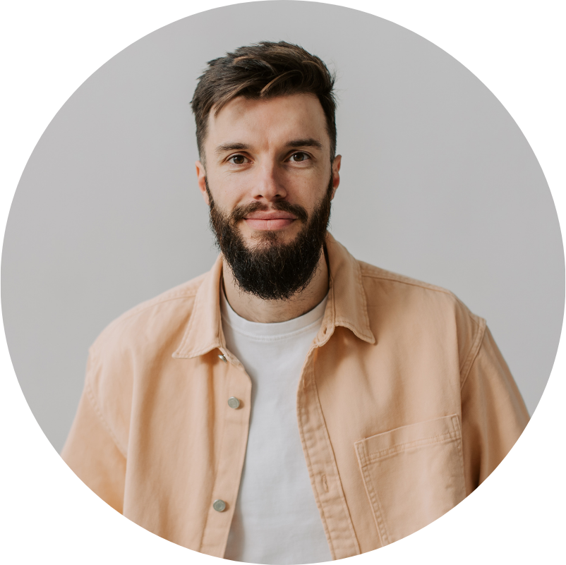 Man with dark hair and beard smiles; wears light orange jacket and white t-shirt, neutral background.