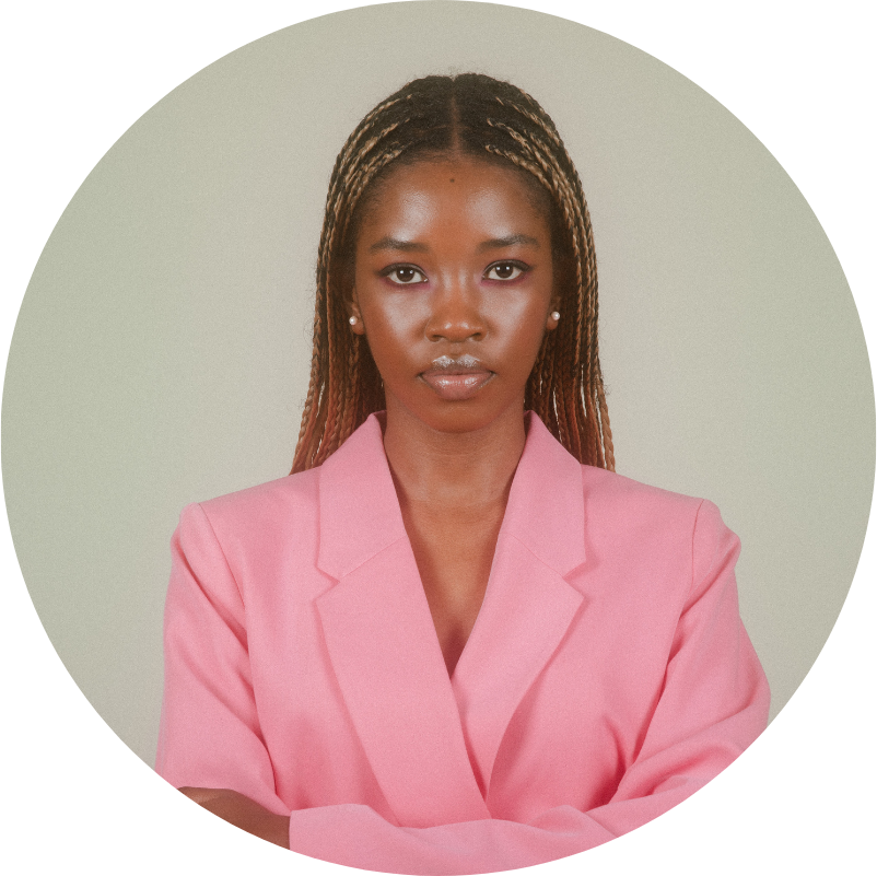 Woman with braided hair wearing a pink blazer, arms crossed, neutral expression.