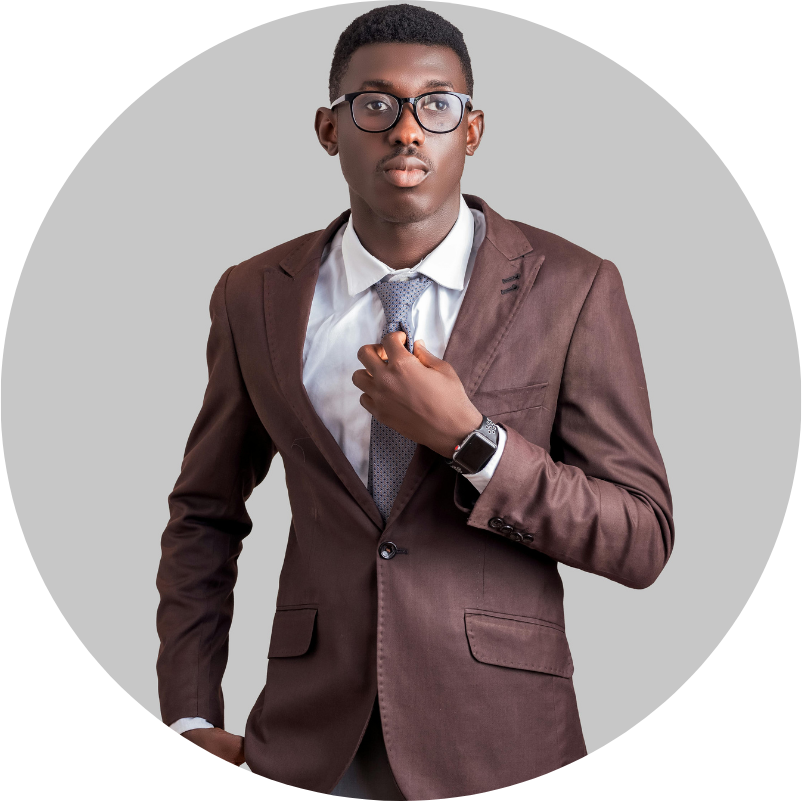 A young Black man in a brown suit and glasses adjusts his tie, standing against a grey background.
