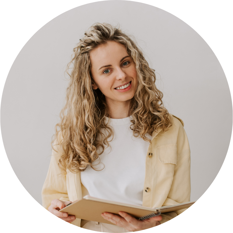 Woman with curly blonde hair smiling, holding a book, wearing a white shirt and yellow jacket.