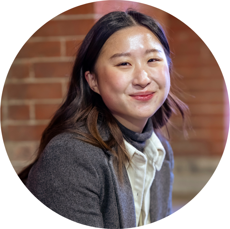 Smiling Asian woman with dark hair, in blazer, against a brick wall.