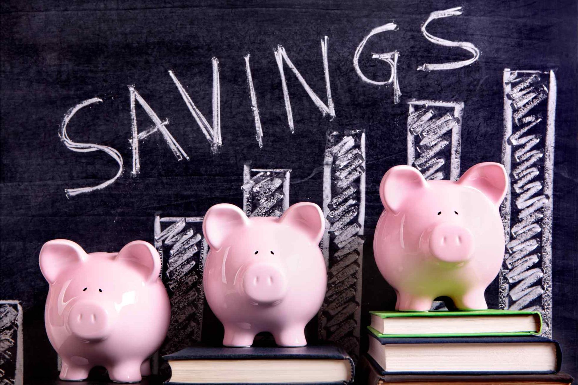 Three pink piggy banks are sitting on top of books in front of a chalkboard that says savings