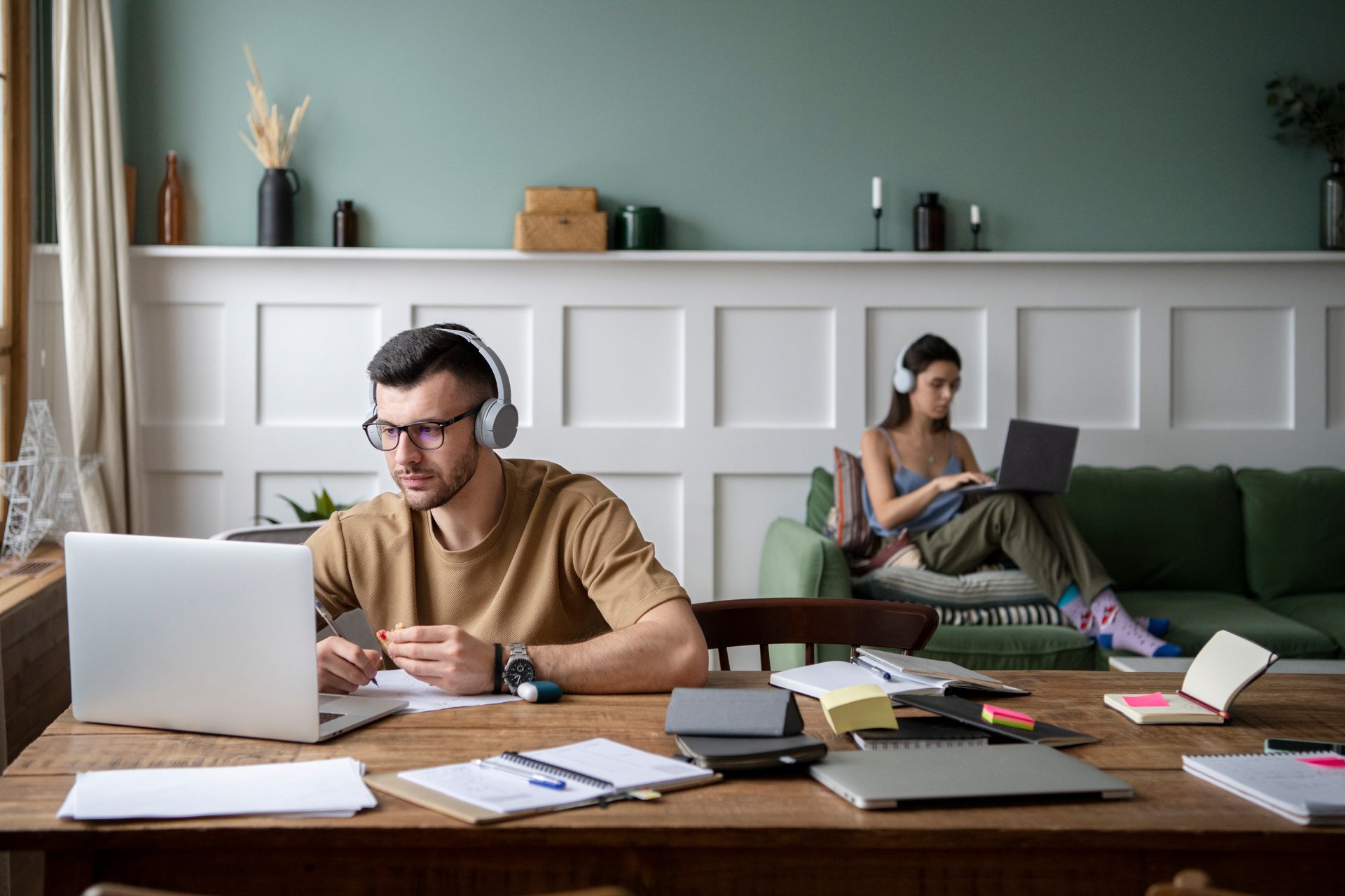 A man is sitting at a table using a laptop while a woman sits on a couch using a laptop.