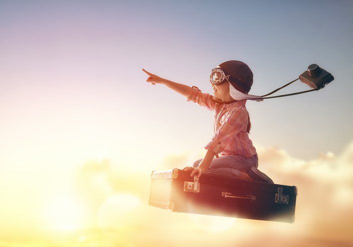 A child dressed like a pilot with a camera rides a briefcase in the clouds and points forward.