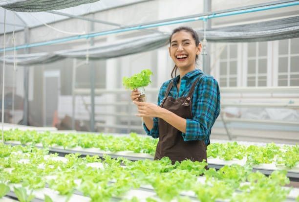 A woman in a lab coat is standing in front of a wall of green plants.