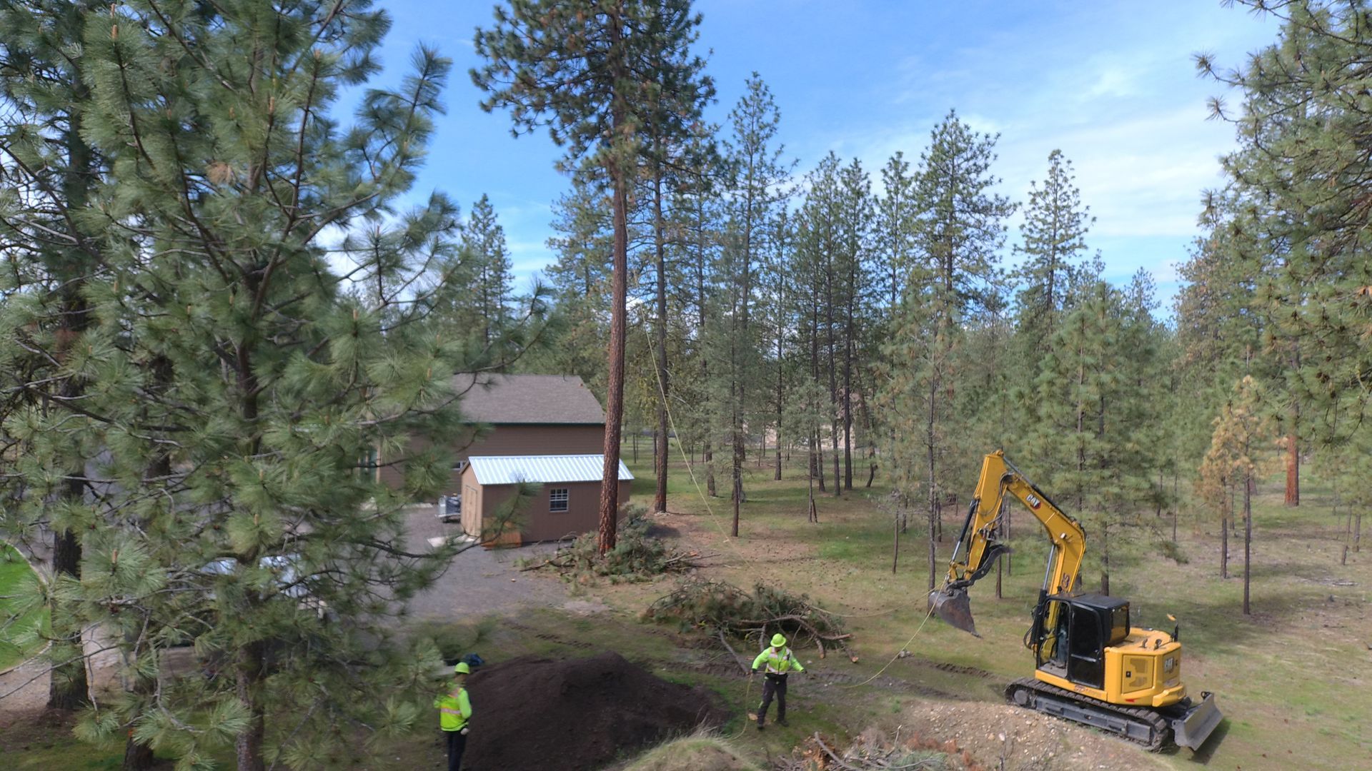 An aerial view of a house in the middle of a forest with a bulldozer in the foreground.