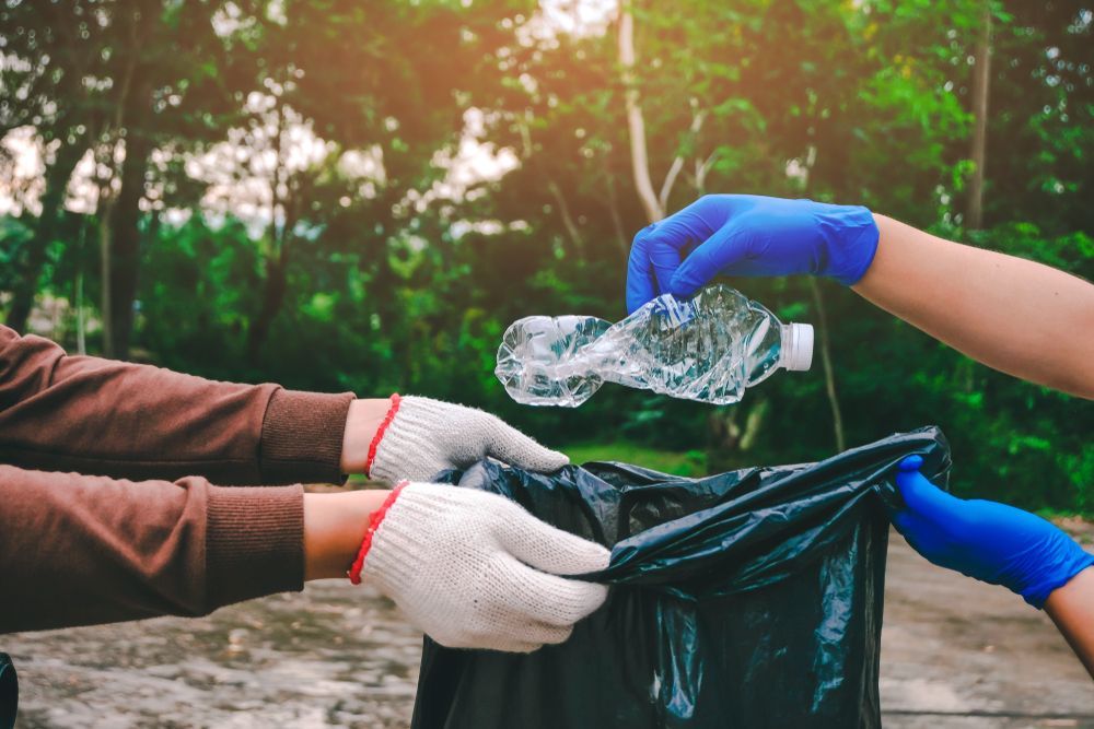 A Person Wearing Blue Gloves Is Putting A Plastic Bottle Into A Trash Bag — Green Solutions Qld Pty Ltd in Mulara, QLD