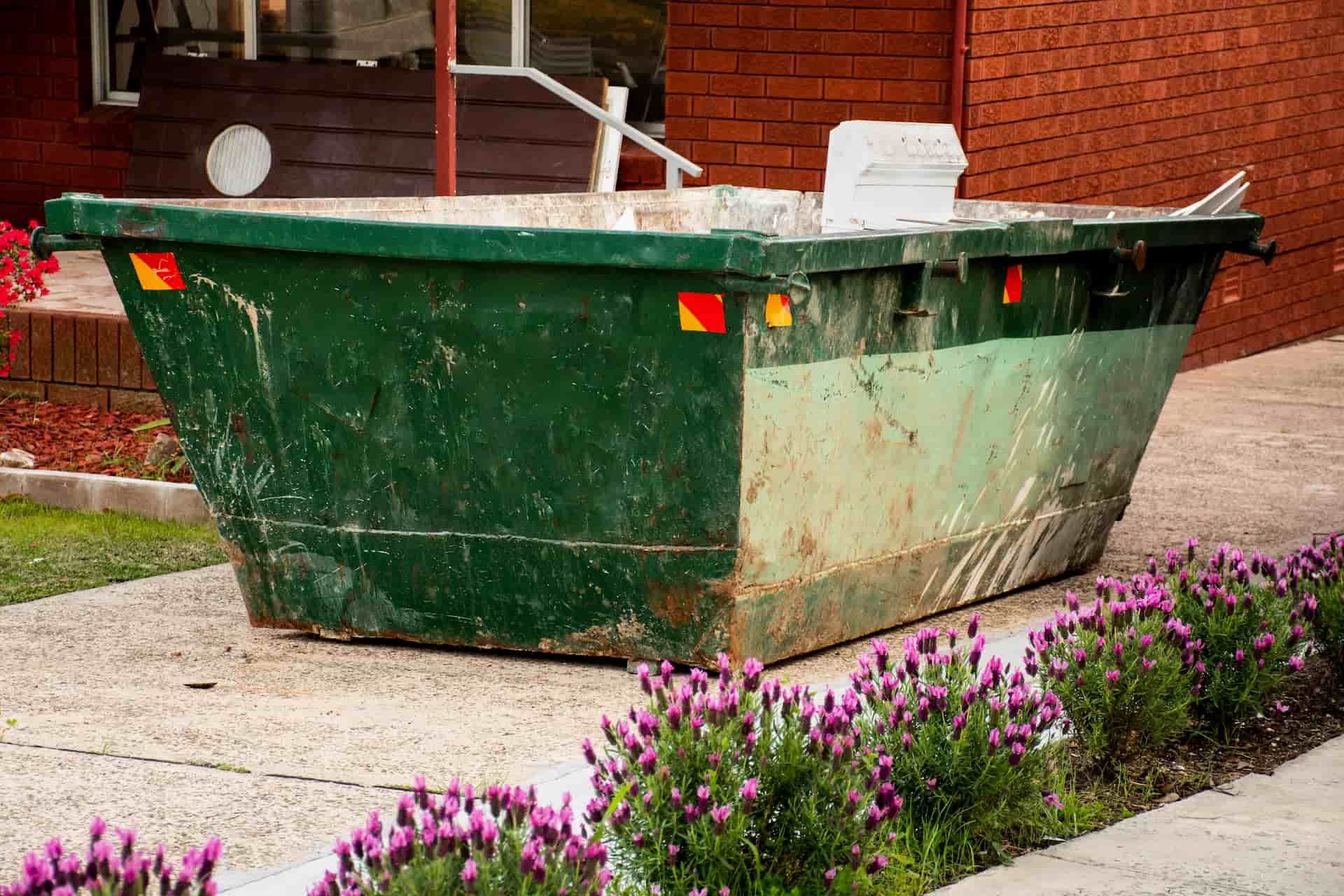 A Green Dumpster Is Sitting On The Sidewalk In Front Of A Brick Building — Green Solutions Qld Pty Ltd in Mulara, QLD