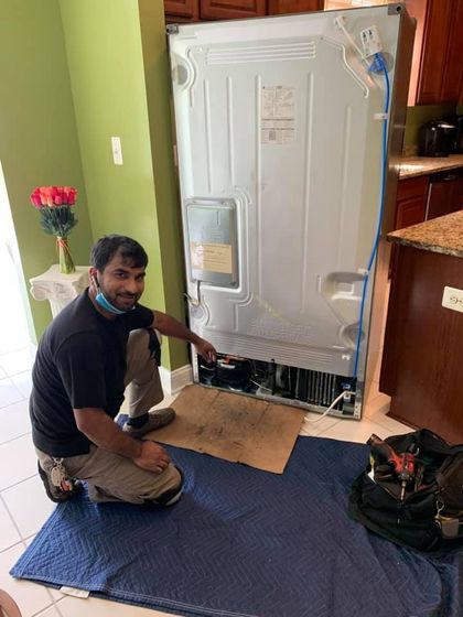 Man kneeling, pointing to the back of a refrigerator. He wears a face mask, and work clothes in a kitchen setting.