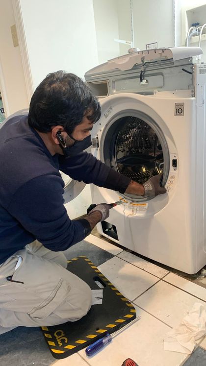 Person repairing a white washing machine, using a screwdriver, inside a bright room.