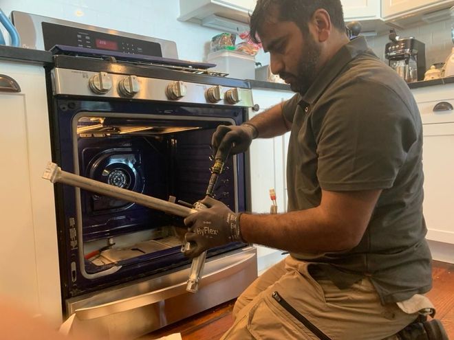 Man in gloves fixing oven, kneeling. Silver oven, light interior, light brown pants, green shirt.