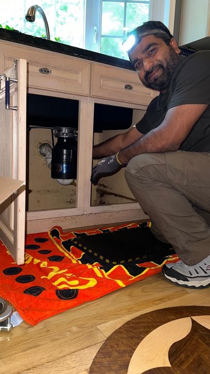 Man kneeling, pointing to the back of a refrigerator. He wears a face mask, and work clothes in a kitchen setting.