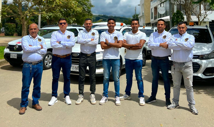 Un grupo de hombres con camisas blancas posan frente a automóviles bajo un cielo nublado.