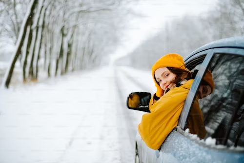 A woman is sticking her head out of a car window on a snowy road.