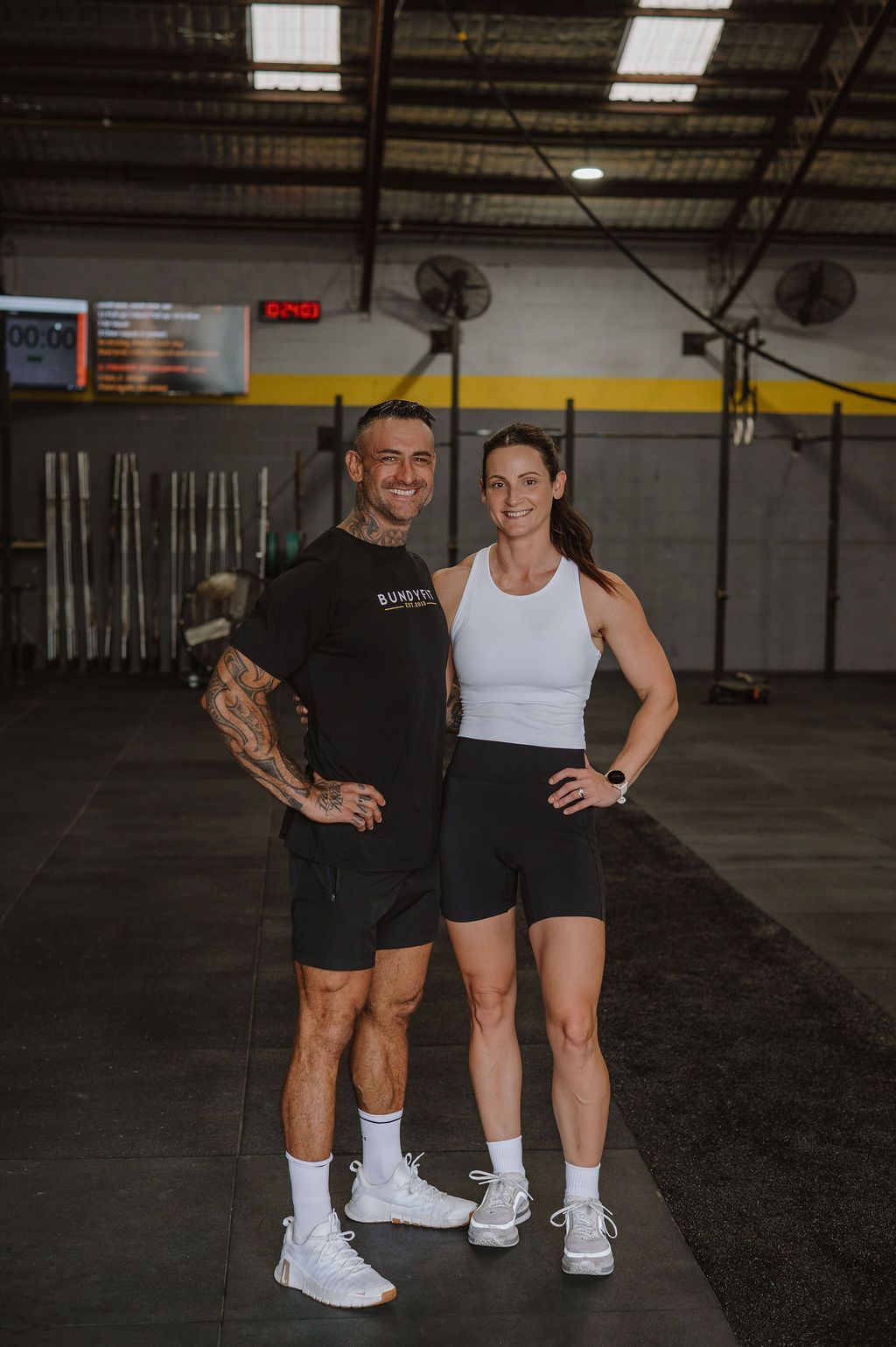 A smiling athletic couple stand together in a gym, wearing workout clothes and sneakers, posing for a photo.