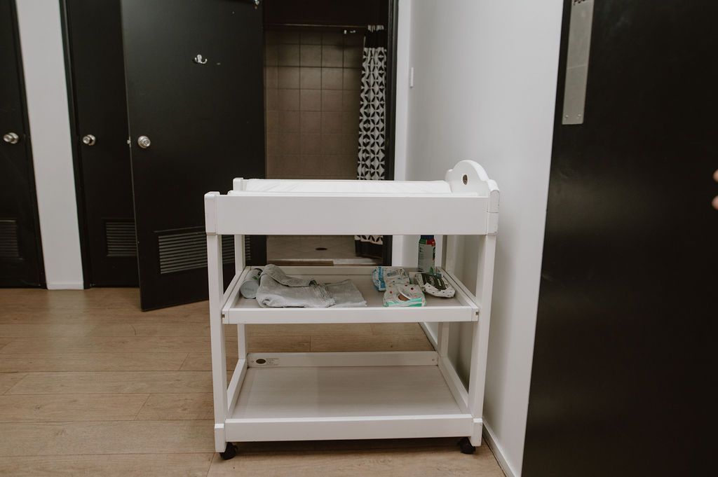A white changing table on wheels stands in a room with black doors and a tiled floor.