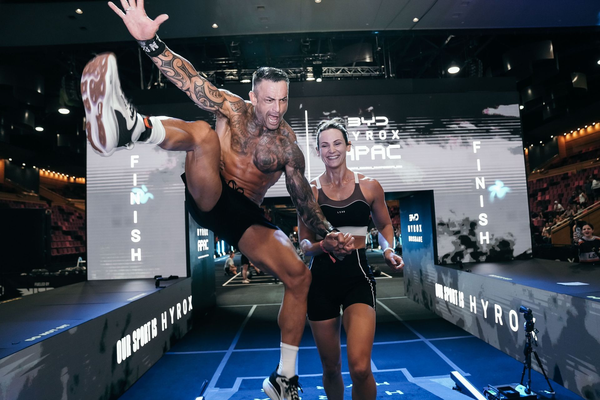 Two people posing on a blue-lit event runway with F45 branding and a large screen behind them
