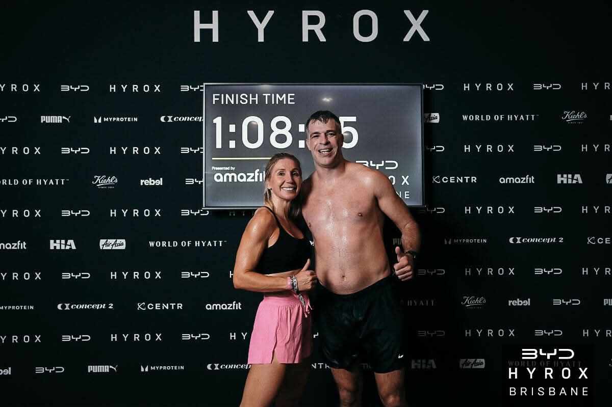 Smiling pair posing at HYROX finish line with 1:08:55 time displayed on screen