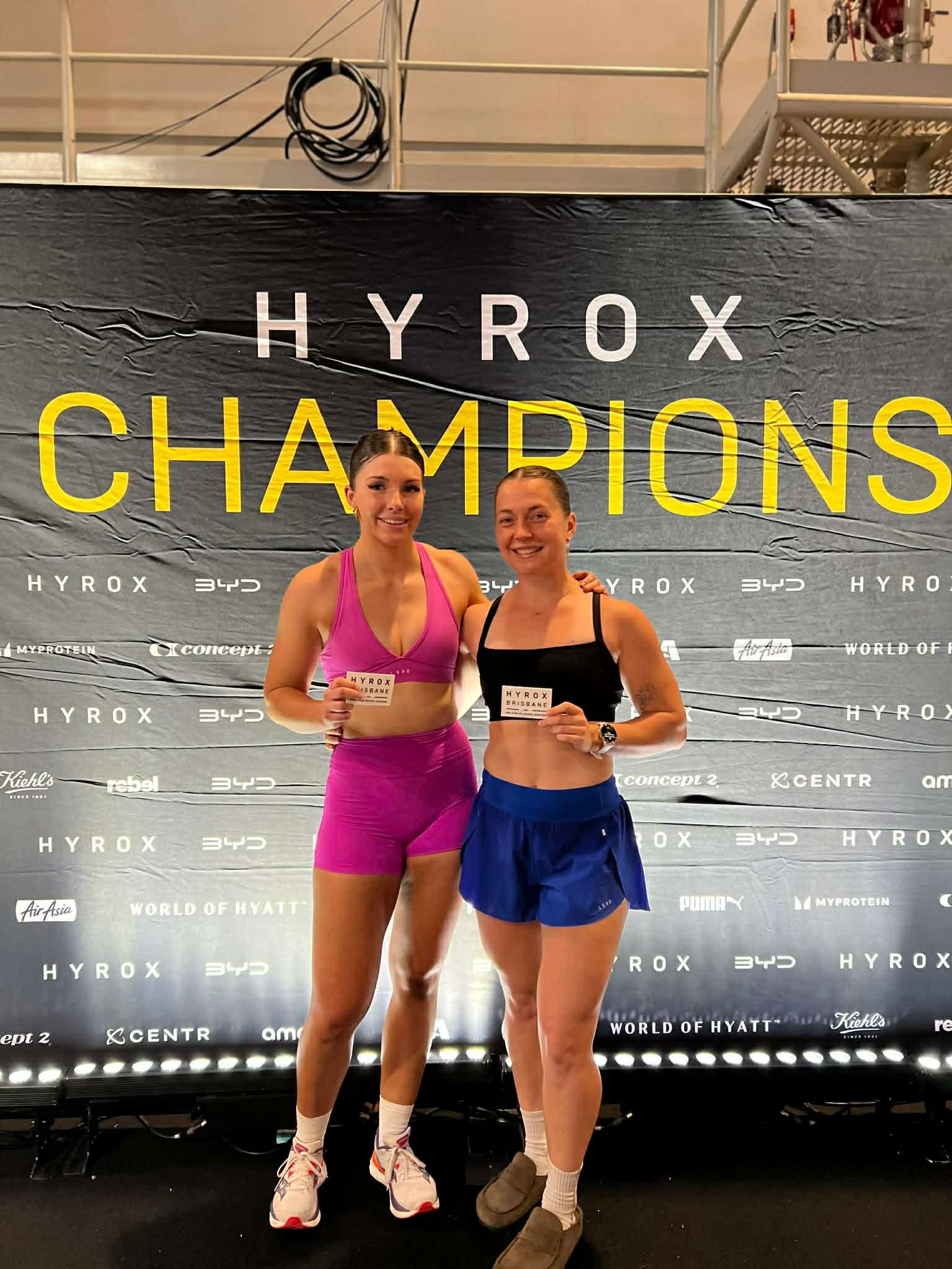 Two athletes pose in front of a “HYROX CHAMPIONS” backdrop, holding medals and smiling.