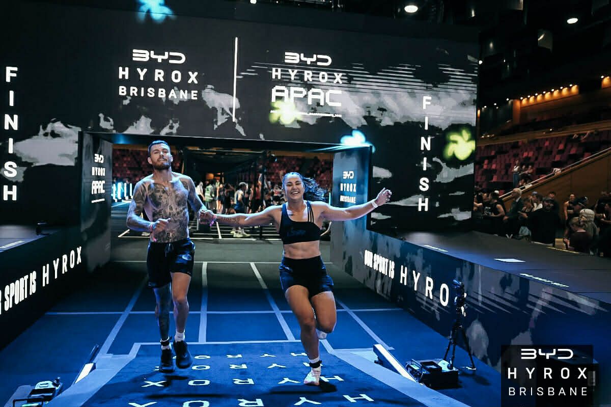 Two athletes sprint toward the finish on a blue HYROX race track indoors