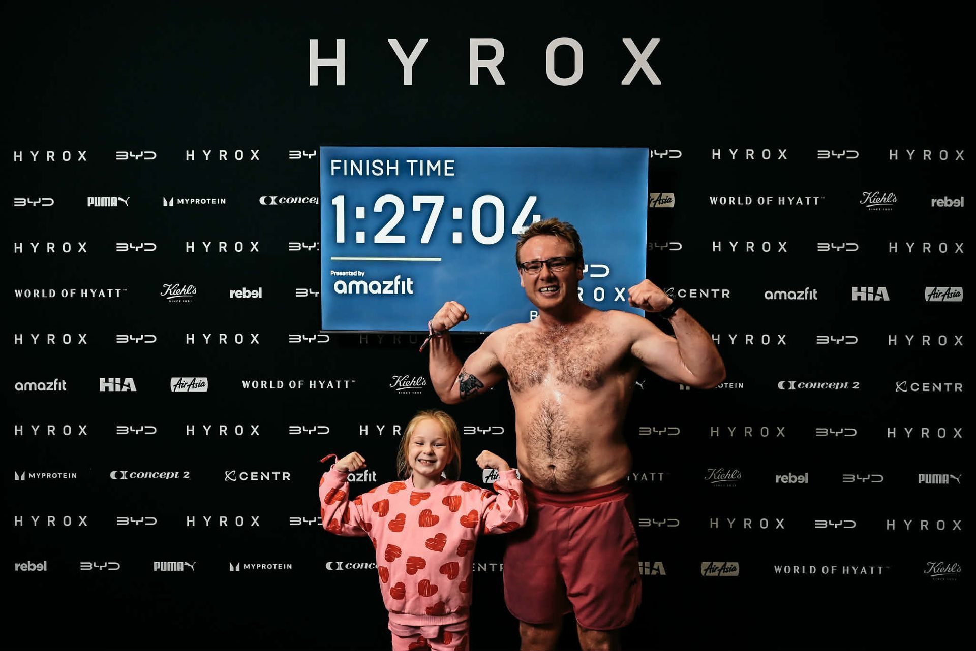 Man and child posing at HYROX finish line with a digital timer reading 1:27:04
