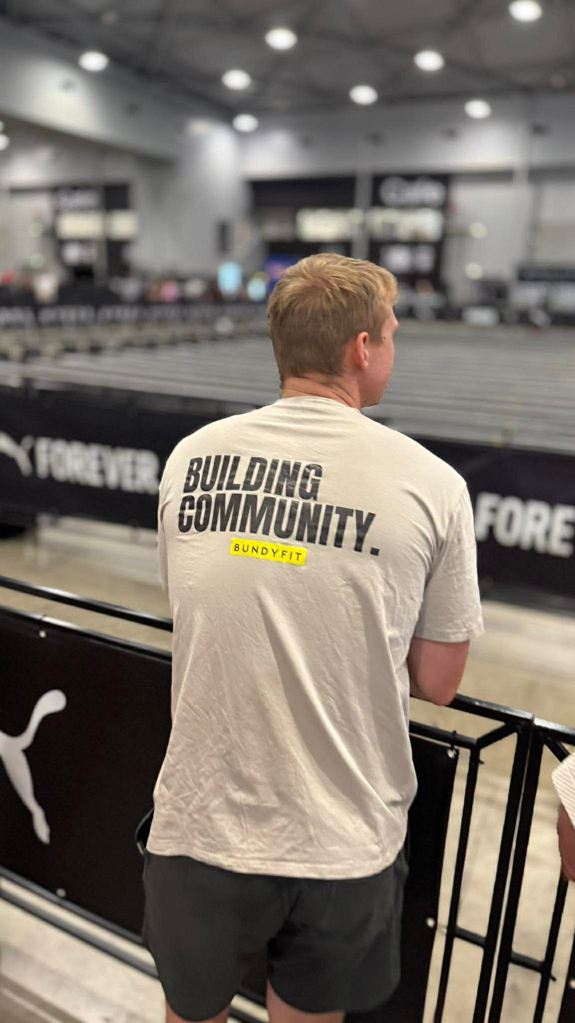 Man in gray T-shirt stands behind a barrier at an indoor sports arena.