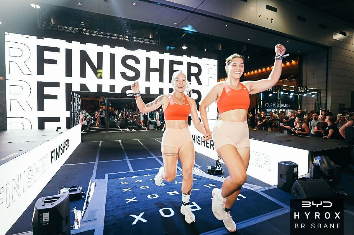 Two runners in orange sportswear cross a finish line at an indoor race event
