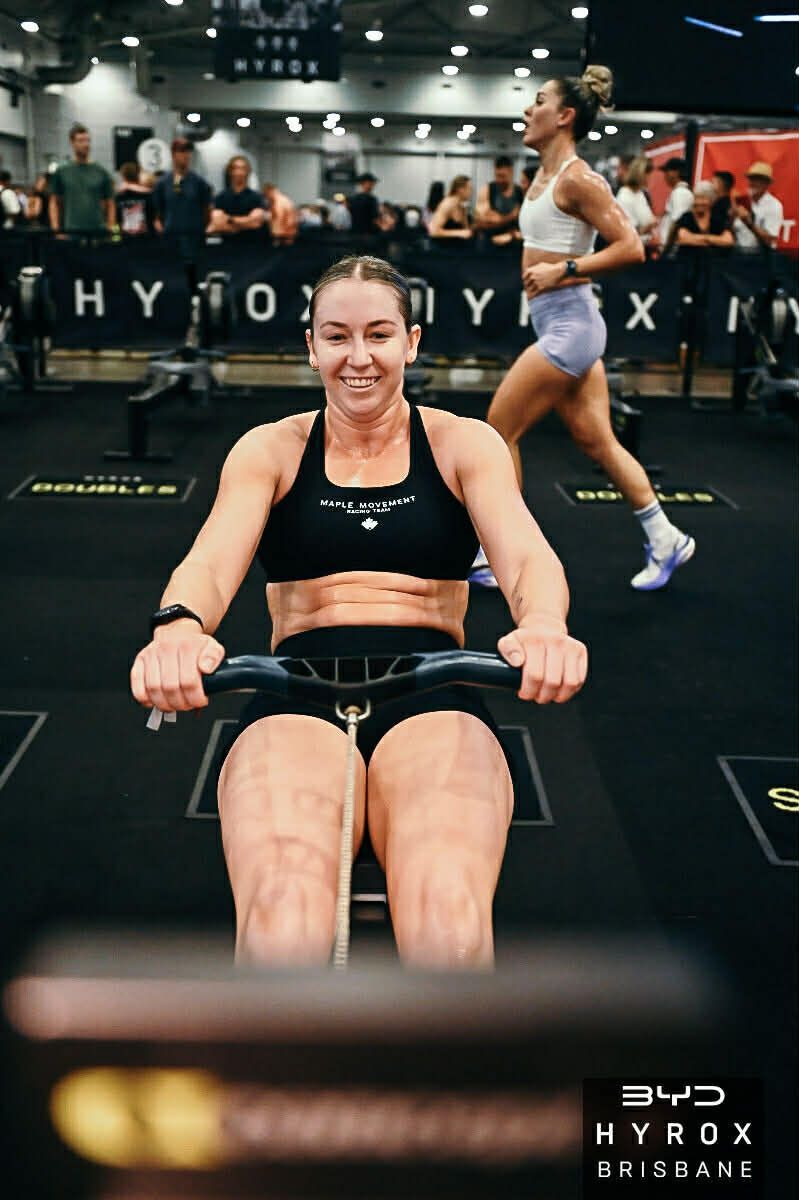 Woman rowing on an erg in a gym during a HYROX event, smiling and wearing black workout gear