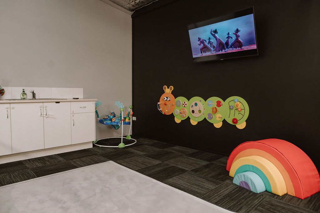 A daycare room with a black wall, a caterpillar decal, a rainbow-shaped foam climber, a TV, and a white cabinet.
