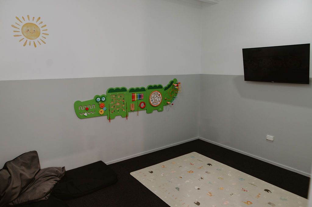 A child's playroom with a light-colored play mat, a green crocodile wall toy, a wall-mounted TV, and a beanbag chair.