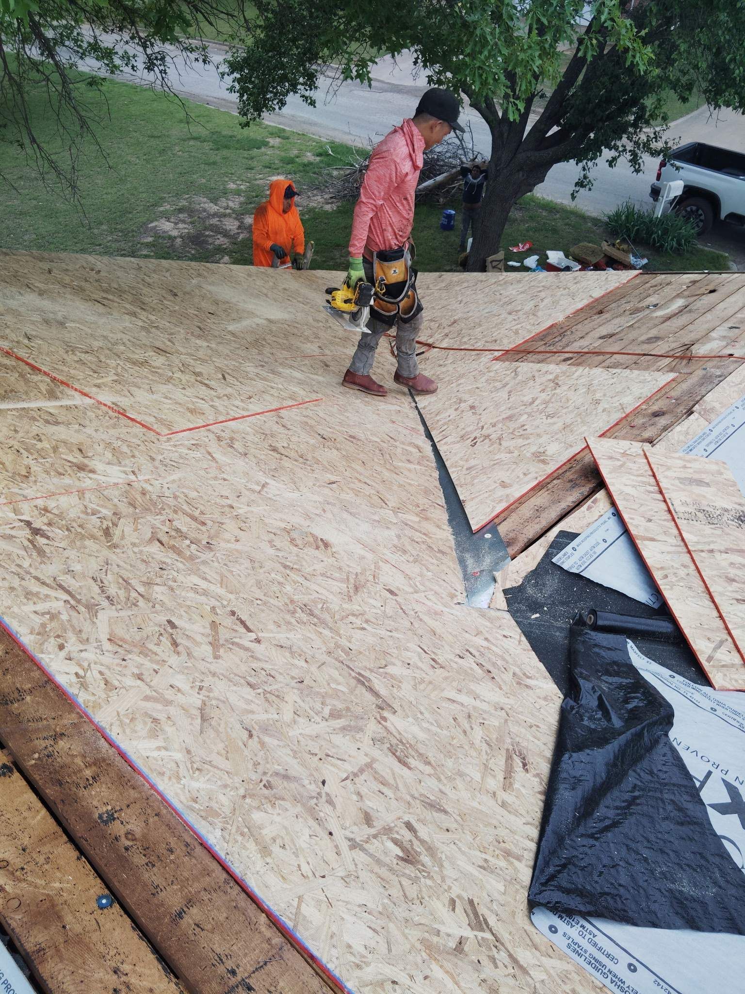 A roofer works on a roof, with tools and another worker in orange visible.