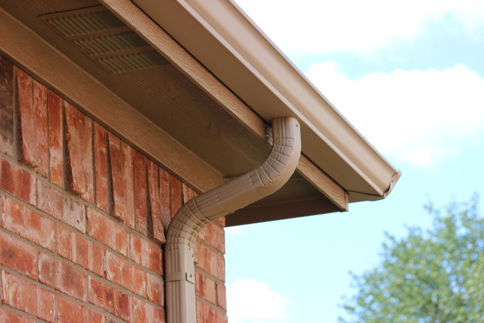 Tan gutter and downspout on a brick building with a blue sky background.
