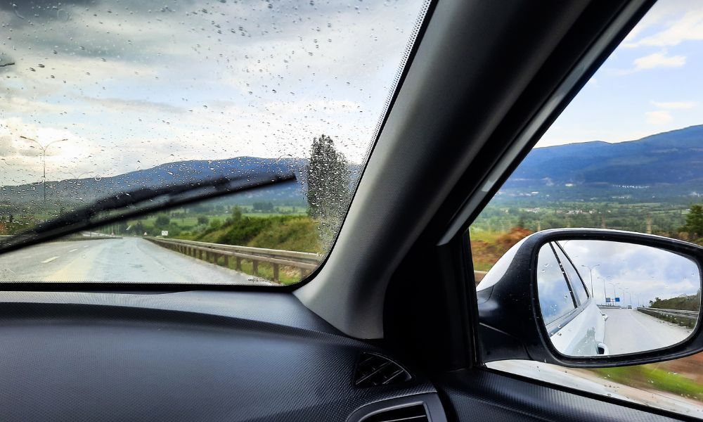 Inside car, wipers clearing rain from windshield; view of road, mountains, side mirror reflecting car.
