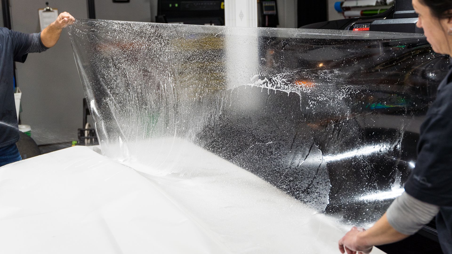 Two people applying clear protective film to a car surface, indoors.