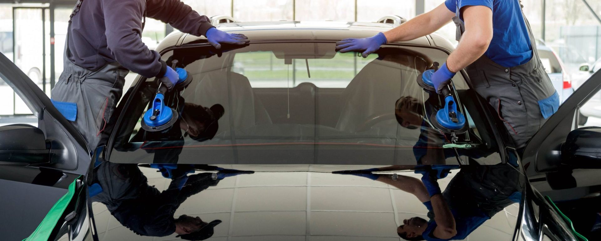 Two workers install a car window. They wear gloves and use suction cups. Black car is in a shop.