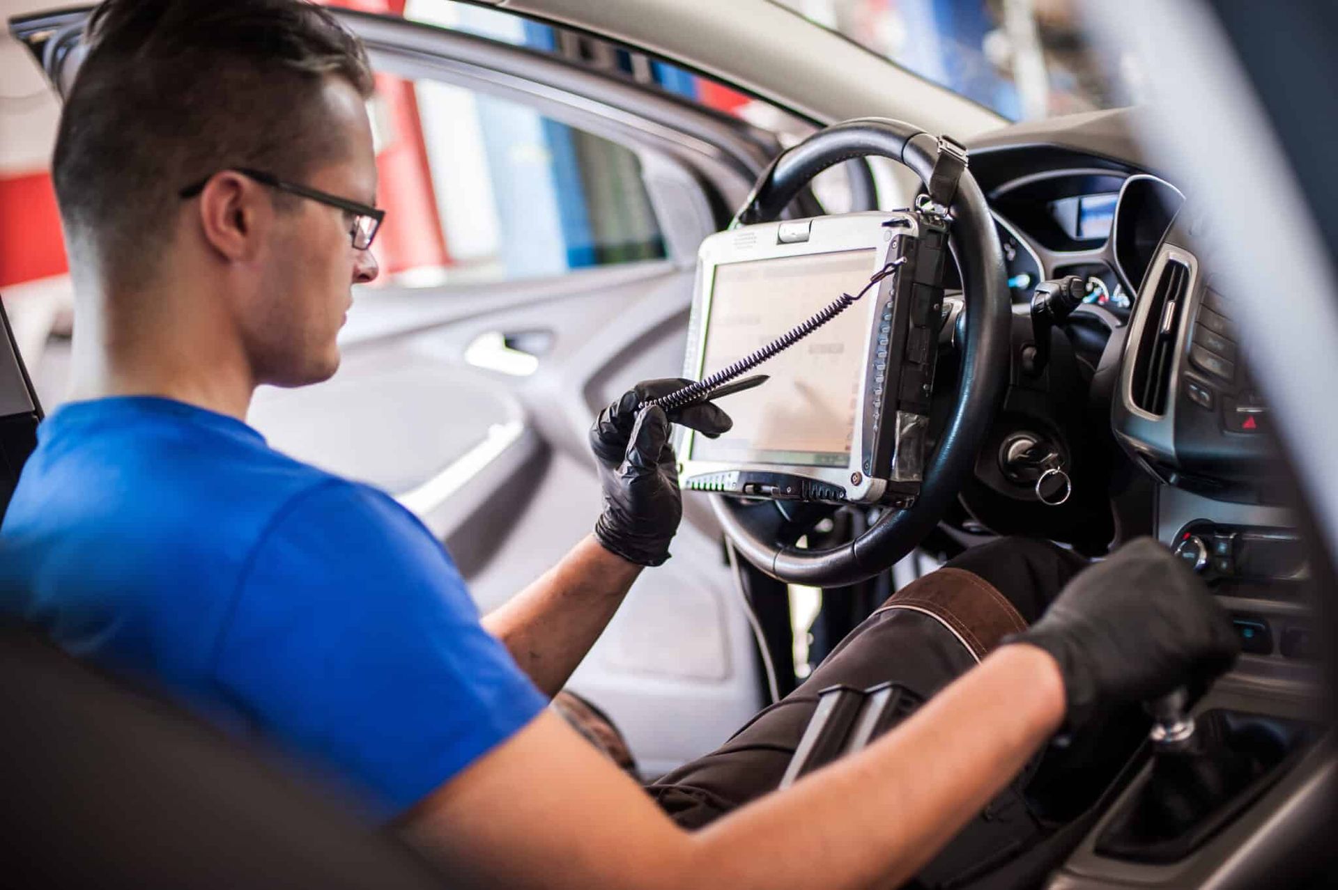 Mechanic using a diagnostic tablet inside a car, wearing glasses and gloves.