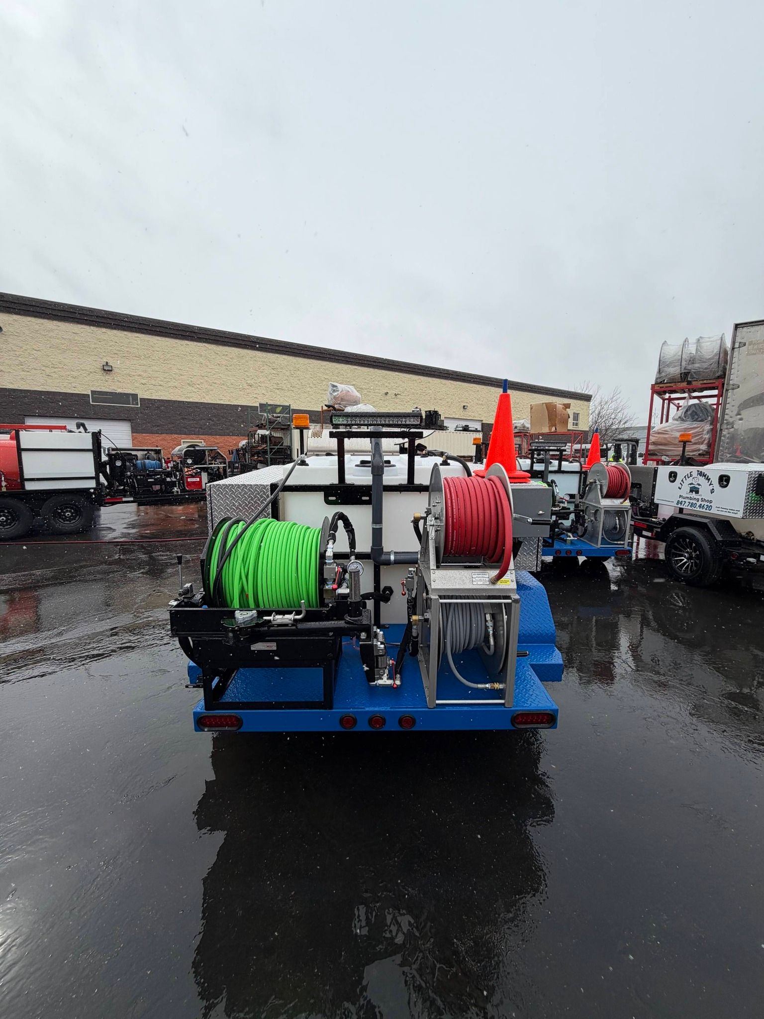 Blue utility trailer with green and red hose reels parked on wet pavement in an industrial lot