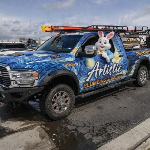 Blue Artistic Plumbing & Drains pickup with a large bunny decal, parked in a lot under cloudy skies