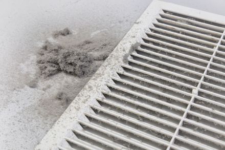 A dusty white wall vent with a pile of grey dust and lint accumulated next to it on the floor.