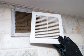 A gloved hand holds a white air vent cover removed from a wall, revealing a dark, clogged vent underneath.