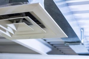A white square ceiling air vent mounted on a metal duct system against a ribbed industrial ceiling.