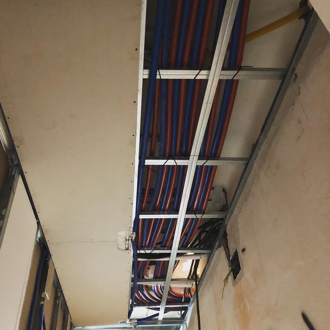 Vista interna di una stanza in costruzione; soffitto a vista con tubi blu e rossi, cartongesso e struttura metallica.